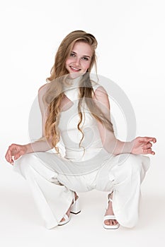 Beautiful blonde woman dressed in white jumpsuit sitting squatting position and looking at camera