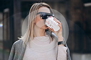 Beautiful blonde woman drinking coffee on street.Portrait of stylish smiling business woman in dark trousers, creamy