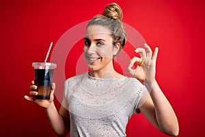 Beautiful blonde woman drinking cola fizzy beverage to refreshment over red background doing ok sign with fingers, excellent