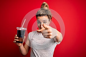 Beautiful blonde woman drinking cola fizzy beverage to refreshment over red background pointing with finger to the camera and to