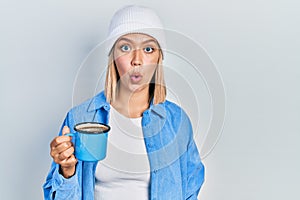 Beautiful blonde woman drinking a cup of coffee scared and amazed with open mouth for surprise, disbelief face