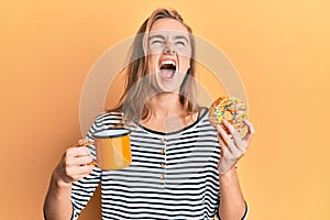 Beautiful blonde woman eating doughnut and drinking coffee angry and mad screaming frustrated and furious, shouting with anger