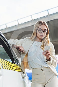 beautiful blonde woman in eyeglasses leaning at taxi cab and using smartphone