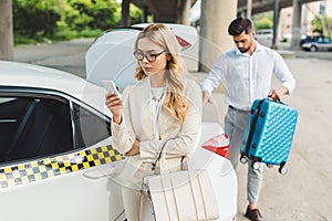 beautiful blonde woman in eyeglasses using smartphone while man putting suitcase in trunk of taxi car
