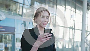 A beautiful blonde woman in a formal black outfit stands by the airport entrance, uses her cell phone, gets the text