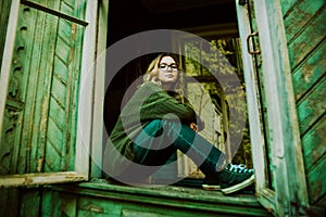 A beautiful blonde woman in a green vintage sweater and jeans is sitting on the windowsill of a wooden abandoned house with broken