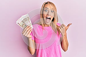 Beautiful blonde woman holding 100 russian ruble banknotes pointing thumb up to the side smiling happy with open mouth