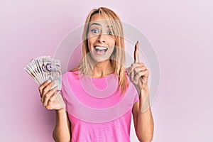 Beautiful blonde woman holding 2 singapore dollars banknotes smiling with an idea or question pointing finger with happy face,