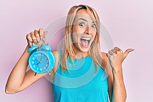 Beautiful blonde woman holding alarm clock pointing thumb up to the side smiling happy with open mouth