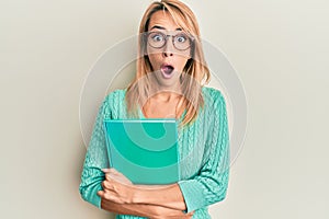 Beautiful blonde woman holding book wearing glasses afraid and shocked with surprise and amazed expression, fear and excited face