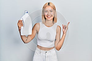 Beautiful blonde woman holding detergent bottle smiling happy pointing with hand and finger to the side