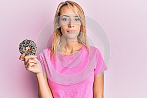 Beautiful blonde woman holding donut thinking attitude and sober expression looking self confident