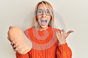 Beautiful blonde woman holding healthy fresh pumpkin pointing thumb up to the side smiling happy with open mouth