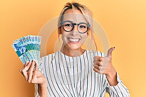 Beautiful blonde woman holding hong kong 20 dollars banknotes smiling happy and positive, thumb up doing excellent and approval