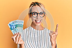 Beautiful blonde woman holding hong kong 20 dollars banknotes smiling with an idea or question pointing finger with happy face,