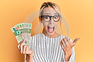 Beautiful blonde woman holding hong kong 50 dollars banknotes pointing thumb up to the side smiling happy with open mouth