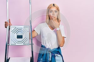 Beautiful blonde woman holding ladder covering mouth with hand, shocked and afraid for mistake