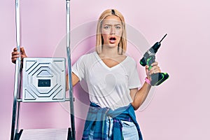 Beautiful blonde woman holding ladder holding screwdriver in shock face, looking skeptical and sarcastic, surprised with open