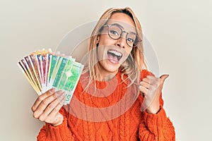 Beautiful blonde woman holding norwegian krone banknotes pointing thumb up to the side smiling happy with open mouth