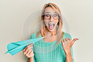 Beautiful blonde woman holding paper airplane pointing thumb up to the side smiling happy with open mouth