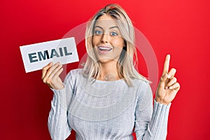 Beautiful blonde woman holding paper with email address smiling with an idea or question pointing finger with happy face, number