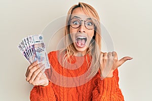 Beautiful blonde woman holding russian ruble banknotes pointing thumb up to the side smiling happy with open mouth