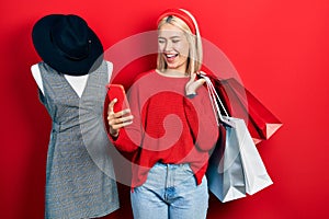 Beautiful blonde woman holding shopping bags and smartphone smiling and laughing hard out loud because funny crazy joke
