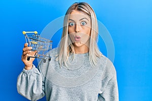 Beautiful blonde woman holding small supermarket shopping cart scared and amazed with open mouth for surprise, disbelief face