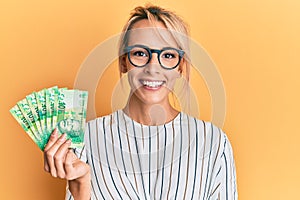 Beautiful blonde woman holding south african 10 rand banknotes looking positive and happy standing and smiling with a confident