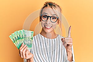 Beautiful blonde woman holding south african 10 rand banknotes smiling with an idea or question pointing finger with happy face,