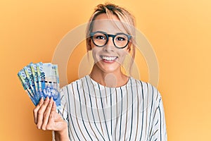 Beautiful blonde woman holding south african 100 rand banknotes looking positive and happy standing and smiling with a confident