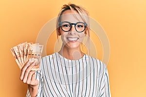 Beautiful blonde woman holding south african 20 rand banknotes looking positive and happy standing and smiling with a confident