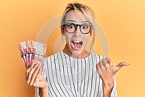 Beautiful blonde woman holding south african 50 rand banknotes pointing thumb up to the side smiling happy with open mouth