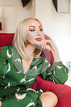 Beautiful blonde woman with makeup in green dress sitting on red velvet sofa