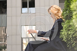 Beautiful blonde woman in navy blue classic smart-casual outfit outdoors near hi-tech business building. She working on her laptop