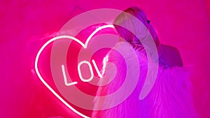 beautiful blonde woman in nightclub with neon pink lights, heart and inscription love on wall