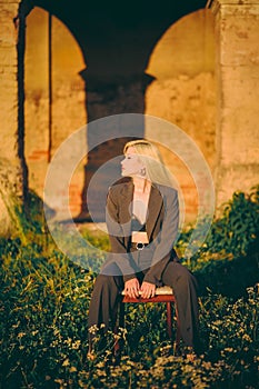 Beautiful blonde woman in an office suit sitting on a chair in nature on the background of an old castle.