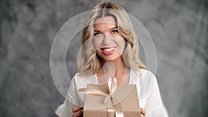 Beautiful blonde woman open desired craft cardboard gift box with bow posing isolated on gray studio