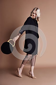 Beautiful blonde woman posing in a black coat with bag on a beige background. Fashion show clothing, woman with perfect figure