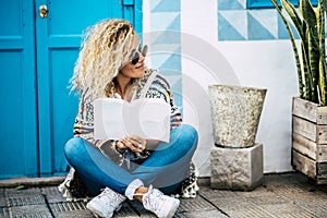 Beautiful blonde woman read a paper book sit down on the ground outside home - blue door and wall background - adult people