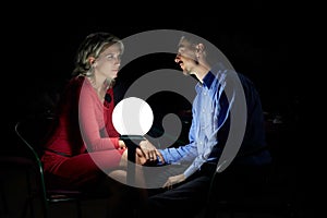 Beautiful blonde woman in red dress, and a man in blue shirt sits together in darkness, next to a ball shaped lamp. Couple dating