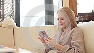 Beautiful blonde woman resting in the lounge area of the bath, looking at photos on smartphone