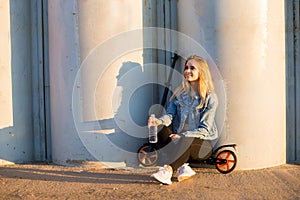 Beautiful blonde woman on scooter near grey columns at sunset holds a water bottle on hand