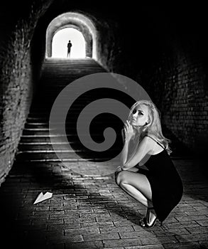 A beautiful blonde woman sits in the old tunnel near the stairs and a paper airplane with the man's silhouette upstairs