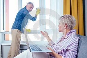 Beautiful blonde woman sitting at sofa and working with laptop,her retired man washing window at living room