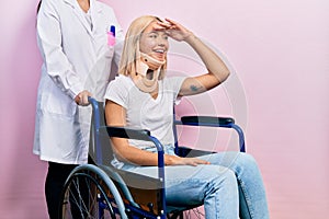 Beautiful blonde woman sitting on wheelchair with collar neck very happy and smiling looking far away with hand over head