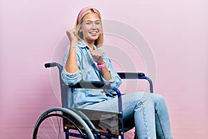 Beautiful blonde woman sitting on wheelchair pointing to the back behind with hand and thumbs up, smiling confident