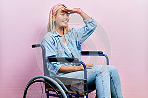Beautiful blonde woman sitting on wheelchair very happy and smiling looking far away with hand over head