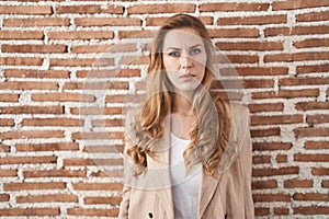 Beautiful blonde woman standing over bricks wall relaxed with serious expression on face