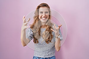 Beautiful blonde woman standing over pink background shouting frustrated with rage, hands trying to strangle, yelling mad